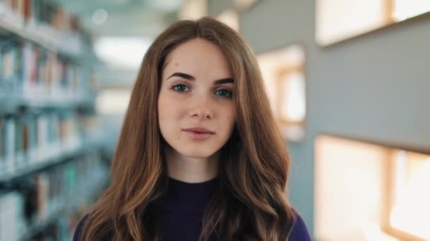 Chica con el pelo largo sonríe y mira directamente a la cámara de pie ante los estantes de la biblioteca — Vídeos de Stock