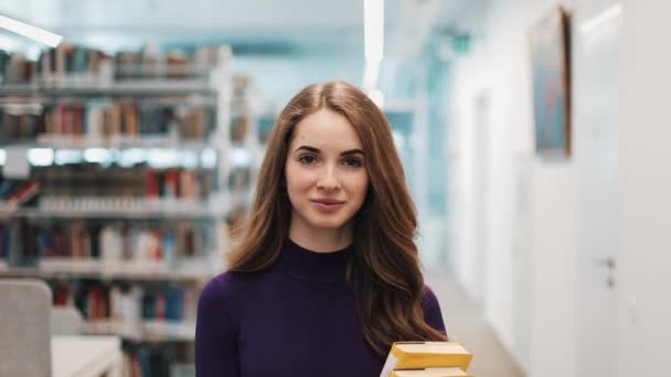 Meisje met lang haar kijkt recht in de camera en wandelingen langs de schappen in de bibliotheek — Stockvideo