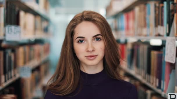 Girl with long hair smiles and looks straight into the camera standing before the shelves in the library — Stock Video