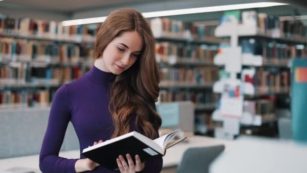 Joven estudiante reflexiva lee un libro de pie en la biblioteca — Vídeos de Stock