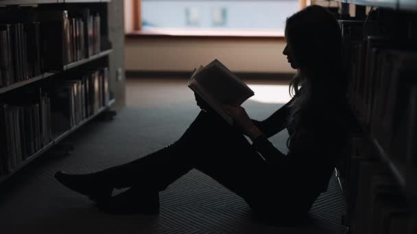 La chica se sienta en el suelo y lee un libro en la biblioteca — Vídeo de stock