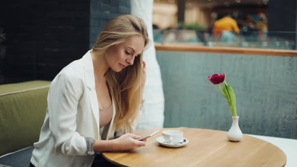 Mooie blonde vrouw leest iets in haar smartphone zitten aan de tafel in café — Stockvideo