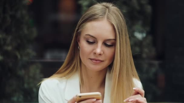 Encantadora mujer rubia lee algo en su teléfono inteligente sentado a la mesa en la cafetería — Vídeo de stock