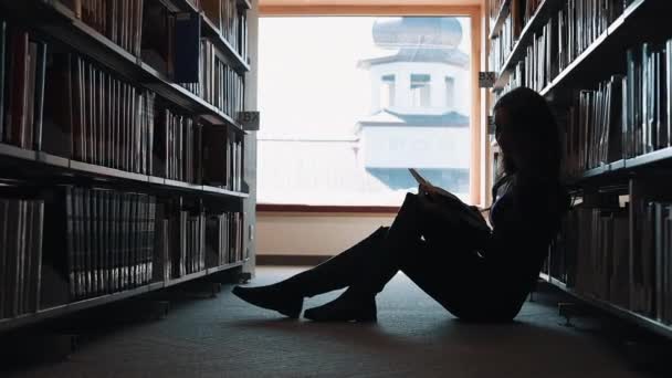 La chica se sienta en el suelo y lee un libro en la biblioteca — Vídeos de Stock