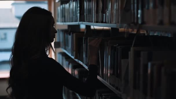 Silhueta de uma menina olhando para os livros antes de uma prateleira na biblioteca — Vídeo de Stock
