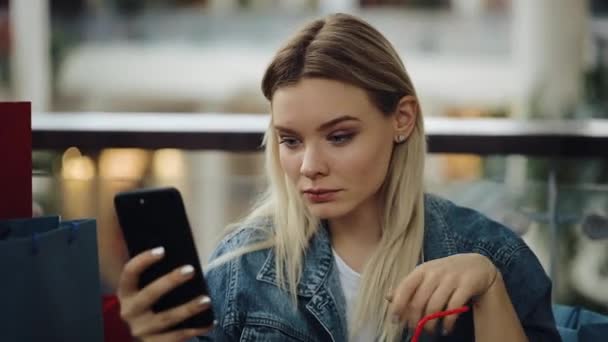 Jolie fille blonde prend selfie sur son téléphone assis dans un café avec des sacs à provisions — Video