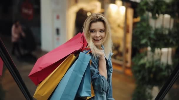 Молодая блондинка в джинсовой куртке улыбается, спускаясь по лестнице в торговом центре — стоковое видео