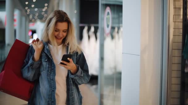 Улыбающаяся блондинка читает что-то в телефоне, гуляя с сумками по торговому центру — стоковое видео