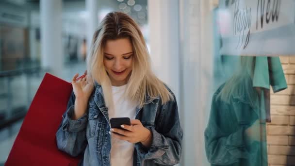 Усміхнена блондинка читає щось у своєму телефоні, ходячи з сумками навколо торгового центру — стокове відео