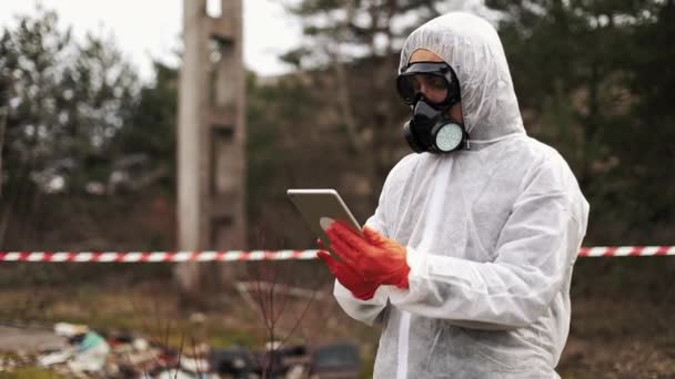 Muž v bio-oblek a plynovou masku trvá o rizicích v jeho postavení tabletu na znečištěné půdy — Stock video