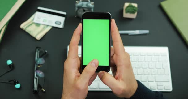 El hombre sostiene un teléfono inteligente negro con pantalla verde sobre una mesa de trabajo y toca algo en él — Vídeos de Stock