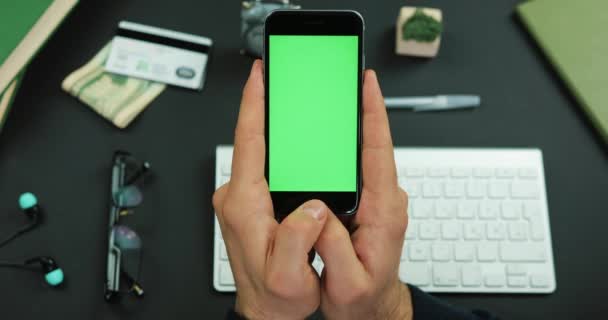 Человек держит черный смартфон с зеленым экраном над рабочим столом и перемещает что-то на нем — стоковое видео