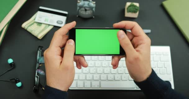 Man holds a black smartphone with green screen over a working table and scrolls something on it — Stock Video