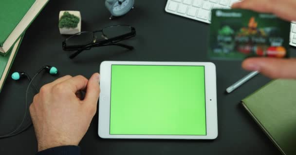 Hombre sostiene una tableta con pantalla verde y escribe la información de la tarjeta de crédito en una mesa con pantalla verde — Vídeo de stock