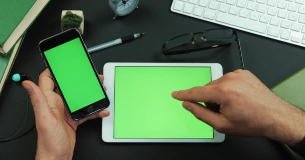 El hombre sostiene el teléfono inteligente con pantalla verde sobre una mesa con pantalla verde y desplaza algo en él — Vídeo de stock