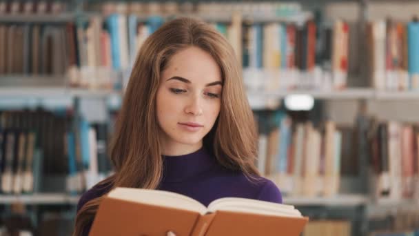 Joven estudiante reflexiva lee un libro de pie en la biblioteca — Vídeos de Stock
