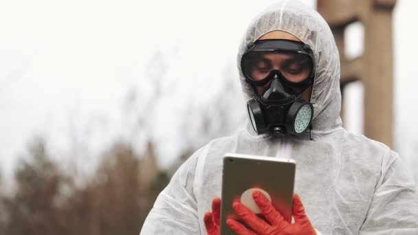 Людина в біо-небезпечному костюмі і газовій масці робить нотатки в планшеті, що стоїть на забрудненій землі — стокове відео