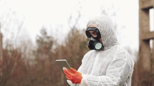 Biyo-tehlike takım elbise ve gaz maskesi alır onun tablet duran kirli karada notlarda erkekte — Stok video