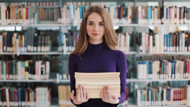 Усміхнена студентка тримає книги, що стоять перед полицями бібліотеки — стокове відео