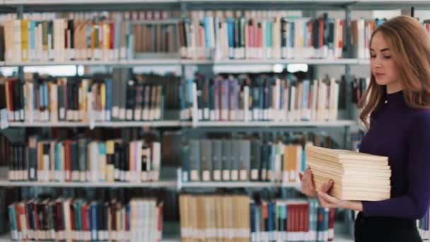 Estudiante sonriente sostiene libros caminando ante los estantes de la biblioteca — Vídeo de stock