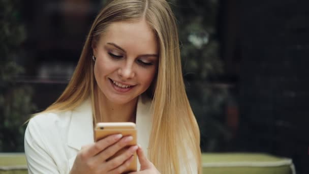 Charmante blonde Frau liest etwas in ihrem Smartphone am Tisch im Café sitzend — Stockvideo