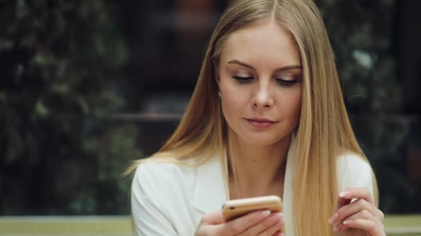 Charmante blonde Frau liest etwas in ihrem Smartphone am Tisch im Café sitzend — Stockvideo