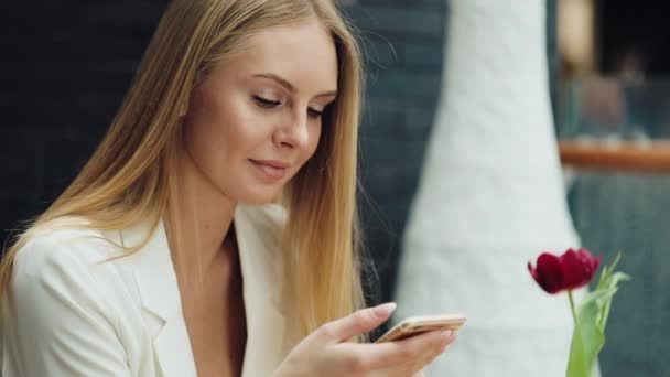 Encantadora mujer rubia lee algo en su teléfono inteligente sentado a la mesa en la cafetería — Vídeos de Stock