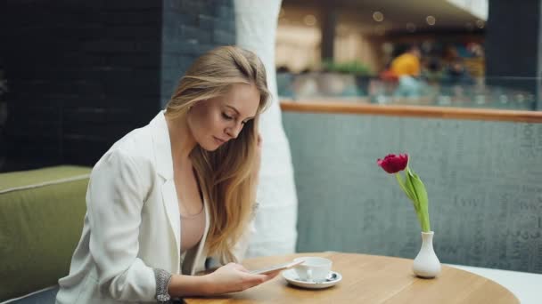 Encantadora mujer rubia lee algo en su teléfono inteligente sentado a la mesa en la cafetería — Vídeos de Stock