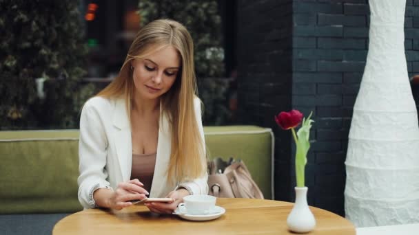 魅力的なブロンドの女性は彼女のスマート フォン カフェでテーブルに座って何かを読み取る — ストック動画