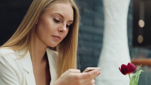 Mooie blonde vrouw leest iets in haar smartphone zitten aan de tafel in café — Stockvideo