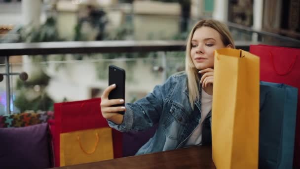 UCRÂNIA, LVIV - FEVEREIRO 2, 2018: Menina loira bonita tira selfie em seu telefone sentado no café com sacos de compras — Vídeo de Stock