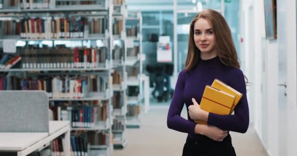 UCRANIA, LVIV - 26 DE MARZO DE 2018: Muchacha con el pelo largo sonríe de pie ante los estantes de la biblioteca — Vídeos de Stock