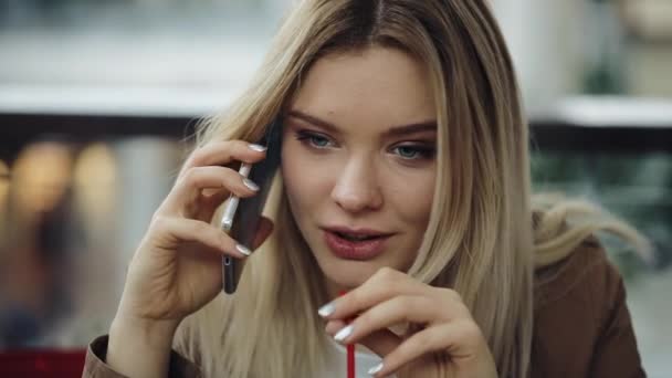 Красивая блондинка разговаривает по телефону, сидит в ресторане и пьет молочный коктейль — стоковое видео