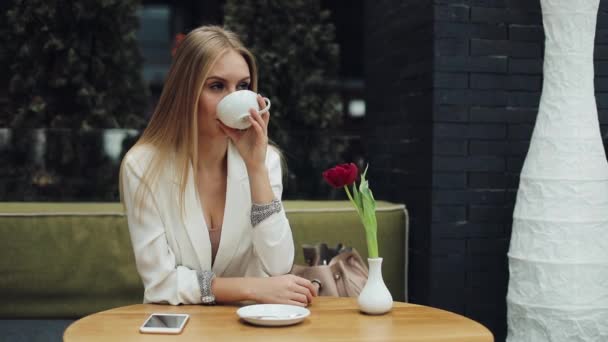 Joven rubia toma una taza de café a sus labios sentados en la mesa en la cafetería — Vídeos de Stock