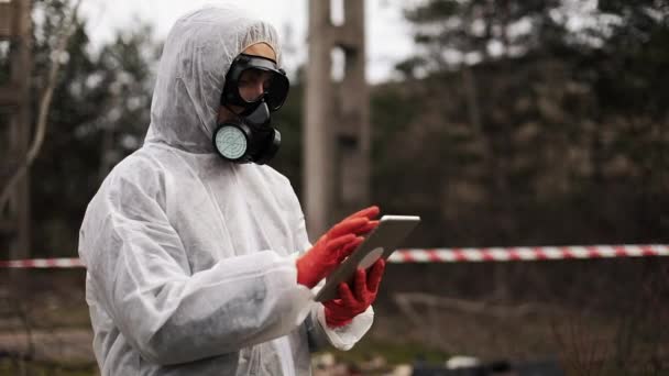 Hombre en traje de bio-peligro y máscara de gas toma notas en su tableta de pie en la tierra contaminada — Vídeos de Stock