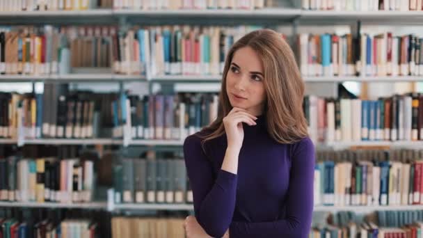 Doordachte jonge vrouw staat voor de schappen in de bibliotheek — Stockvideo