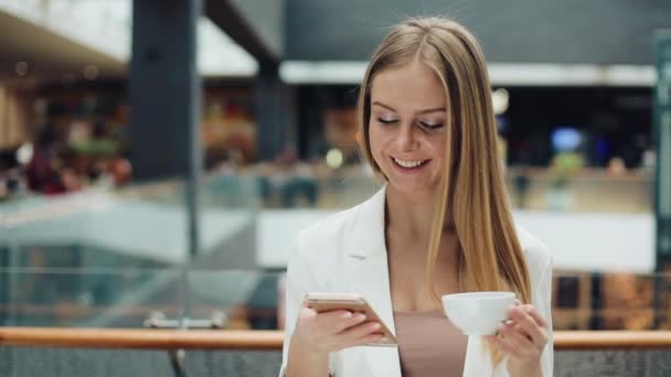 Очаровательная молодая женщина держит смартфон в одной руке, а чашку кофе в другой сидит в кафе — стоковое видео
