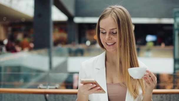 Charming young woman holds smartphone in one hand and cup of coffee in another sitting in the cafe — Stock Video