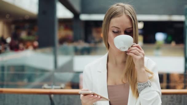 Очаровательная молодая женщина держит смартфон в одной руке, а чашку кофе в другой сидит в кафе — стоковое видео