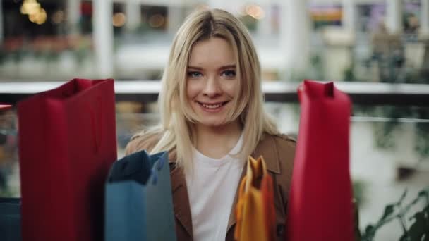 Glada blond kvinna står bakom färgglada shoppingkassar i gallerian — Stockvideo