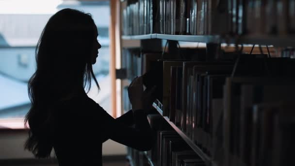 Silueta de una chica mirando los libros antes de un estante en la biblioteca — Vídeos de Stock