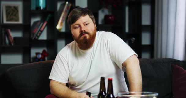 Barbu gros homme boit ours et regarde la télévision avec télécommande à la main, assis à la table avec pop-corn — Video