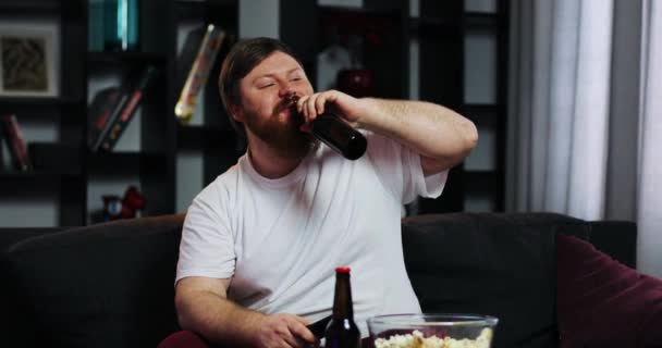 Bärtiger dicker Mann trinkt Bär und schaut Fernsehen mit Fernbedienung in der Hand, sitzt am Tisch mit Popcorn — Stockvideo