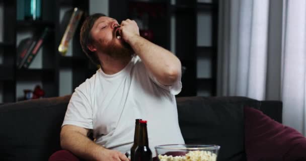 Sorrindo homem gordo com barba assiste TV no quarto e come milho pop — Vídeo de Stock