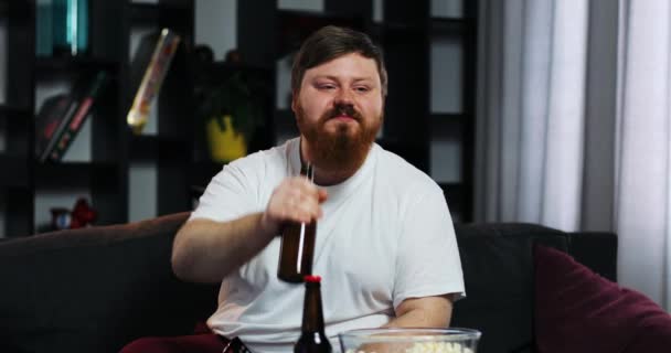 Sonriente hombre gordo con barba mira la televisión en la habitación y bebidas oso — Vídeo de stock