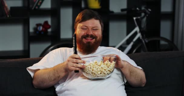 Hombre gordo sonriente con barba mira la televisión en la habitación y come maíz en la mesa con cerveza — Vídeos de Stock