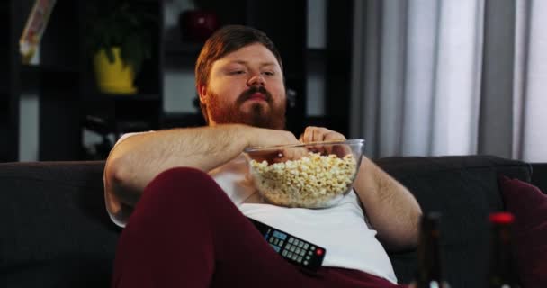 Uomo grasso sorridente con la barba guarda la TV in camera e mangia pop-corn a tavola con la birra — Video Stock