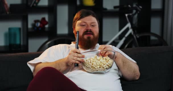 Uomo grasso sorridente con la barba guarda la TV in camera e mangia pop-corn a tavola con la birra — Video Stock