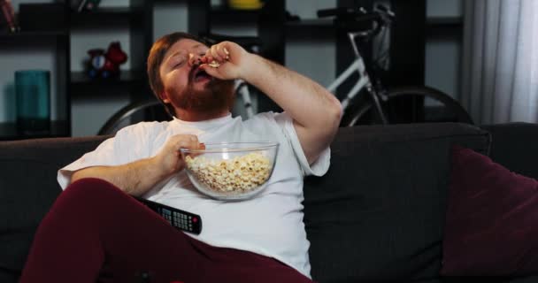 Dikke man met baard glimlachend horloges Tv op de kamer en eet pop-maïs aan tafel met bier — Stockvideo