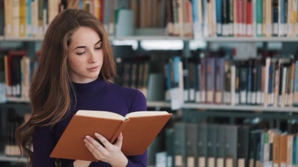 Joven estudiante reflexiva lee un libro de pie en la biblioteca — Vídeo de stock